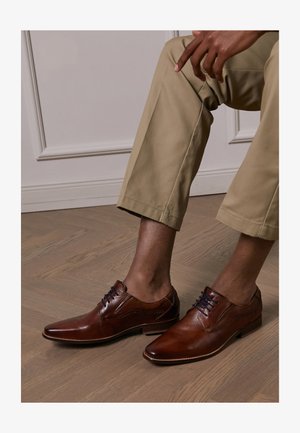 Chaussures habillées en cuir brun avec des pointes pointues, des lacets bleus et des coutures décoratives. Portées avec un pantalon beige clair sur un sol en bois.