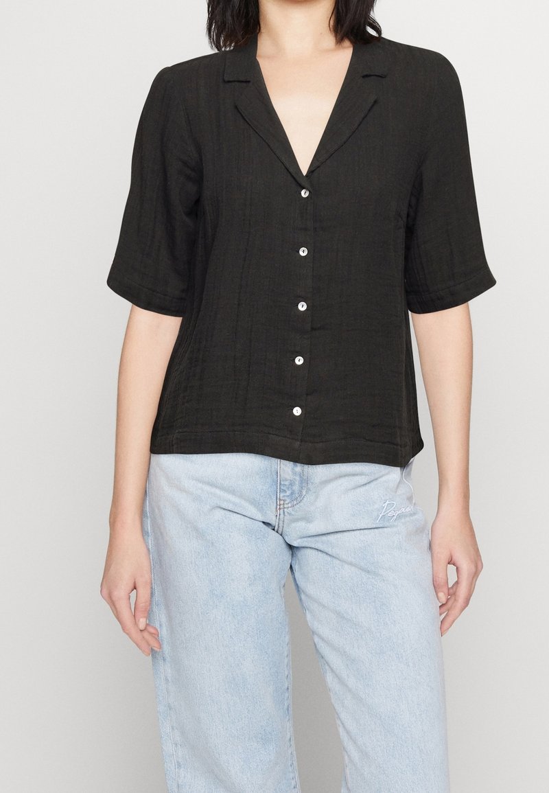Chemise noire à manches courtes avec tissu texturé, col, cinq boutons sur le devant et coupe décontractée. Associée à un jean bleu clair.