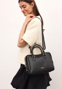 Mustassa nahkakassissa on kaksinkertainen kahva ja säädettävä olkahihna. Varustettu kultasävyisillä tarvikkeilla ja vetoketjulla. Sileä pinta.