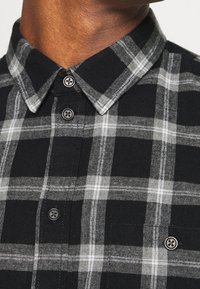 Camicetta a quadri nera e grigia con colletto button-down, dotata di bottoni grigi e una texture morbida, messa in risalto da distintivi motivi a quadri.