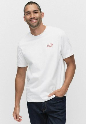 Vit bomull t-shirt med en liten röd logomärke på vänster bröst. Korta ärmar och rundad halsringning. Bärs med mörkblå denimjeans.