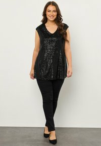 Top nero con paillettes, scollatura a V e maniche corte, abbinato a pantaloni neri attillati e tacchi neri, su uno sfondo neutro.