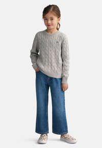 Harmaa cable-knit-neule, jossa on ribbireunat, yhdistettynä leveälahkeisiin sinisiin farkkuihin. Jalkineet ovat vaaleat, joissa on mustia pilkkuja.