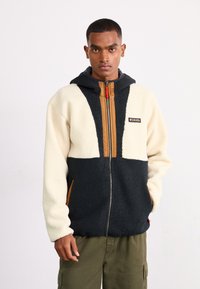 Fleece hættetrøje med cremet overdel og mørkeblå underdel, med en tan lynlås fortil, sidelommer og et logo-patch.