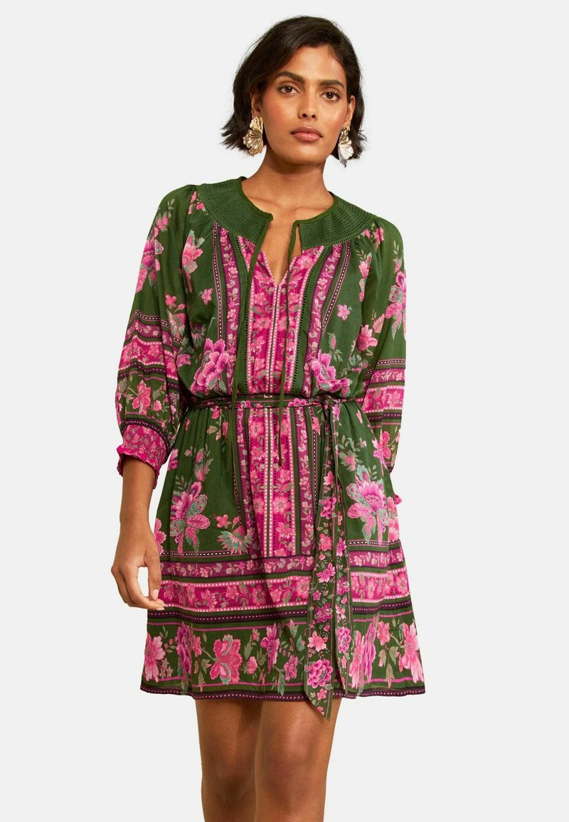 Woman wearing a green and pink floral patterned dress with three-quarter sleeves, a belted waist, and textured round neckline, standing against white background.