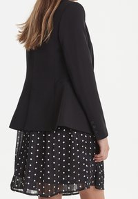 Czarna, dopasowana marynarka z rozszerzanym tyłem, zestawiona z prześwitującą, czarną spódnicą w białe kropki o plisowanej fakturze.