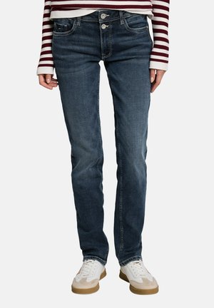 Person trägt dunkelblaue Jeans mit geradem Bein, weiße und beige Sneakers und ein langärmeliges, bordeaux-weiß gestreiftes Oberteil und steht aufrecht.