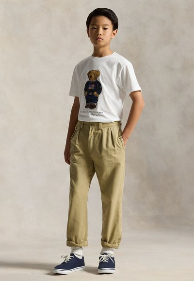 Boy standing with hands in pockets wearing white T-shirt with teddy bear print, beige pants, white socks, and navy blue sneakers.
