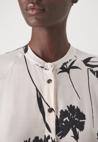 Blusa crema chiara con motivo floreale nero, scollatura rotonda con chiusura a bottoni e texture morbida e liscia.