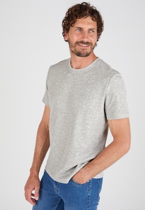 Uomo con capelli ricci che indossa una maglietta a maniche corte grigio chiaro e jeans blu, in piedi con una mano in tasca, che guarda di lato.