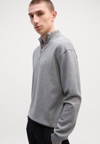 Grauer Reißverschluss-Pullover mit Kragen, gerippten Bündchen und einem dezenten Logodetail auf der Brust. Aus weichem, strukturiertem Stoff gefertigt.