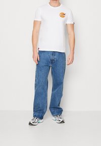 Camiseta blanca de manga corta con un logo redondo de la NASA, combinada con jeans de mezclilla azul de corte holgado. El modelo lleva zapatillas deportivas plateadas.