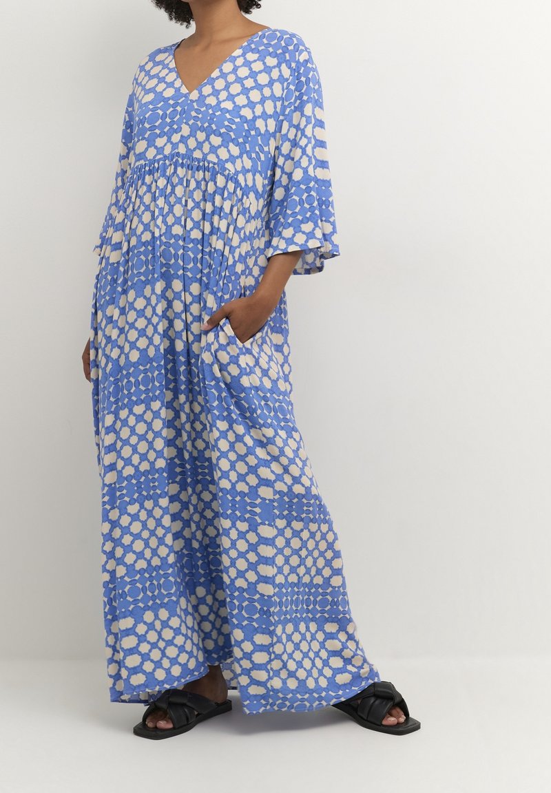 Femme portant une robe maxi ample à motifs bleus et blancs avec un col en V, des manches larges, des poches latérales et des sandales plates noires.