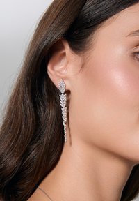 Długie srebrne kolczyki z sylwetką kaskadową, zdobione licznymi kryształowymi akcentami, w zestawieniu z gładką skórą i ciemnymi włosami.