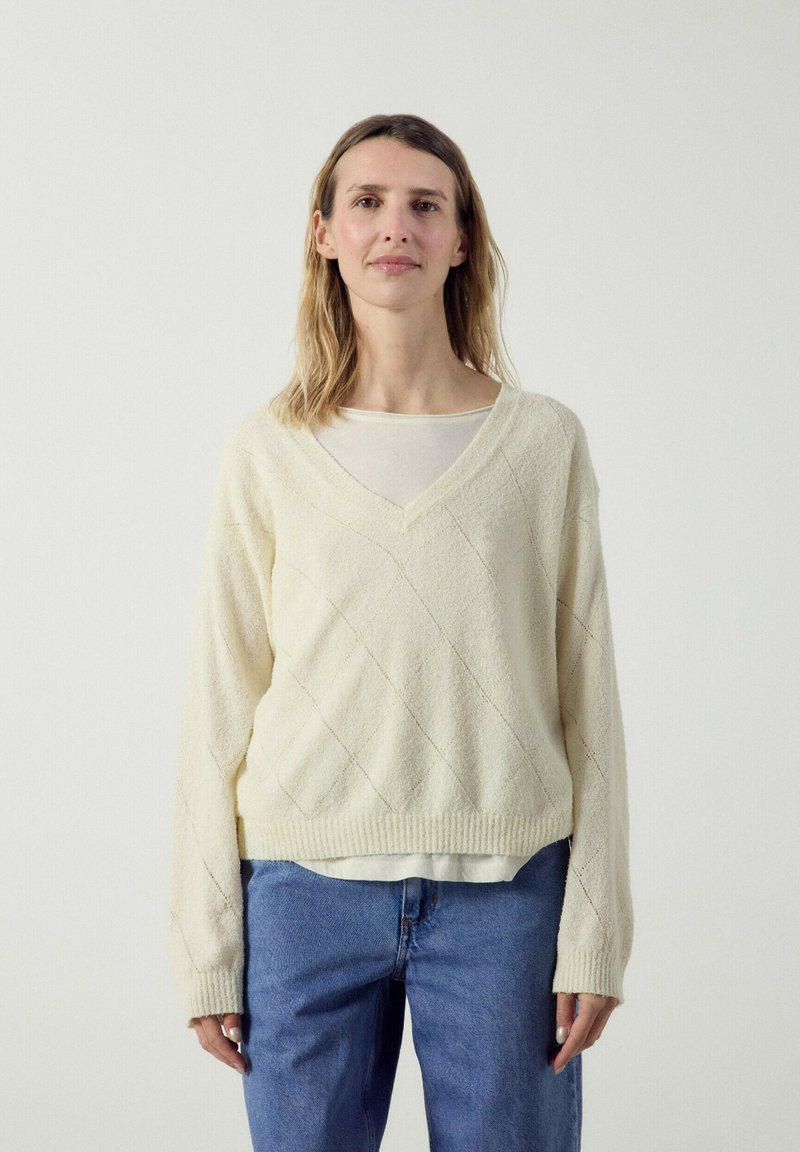Femme aux cheveux mi-longs portant un pull en maille crème à col en V sur un top blanc et un jean bleu, sur un fond clair uni.