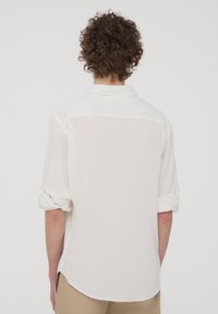 Camicia bianca con maniche arrotolate, caratterizzata da una texture liscia e un orlo curvo, realizzata in tessuto leggero, progettata per un look casual.