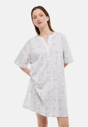 Femme à la peau claire et aux cheveux bruns portant une robe blanche à fleurs, longueur genou, manches courtes et encolure en V fendue.