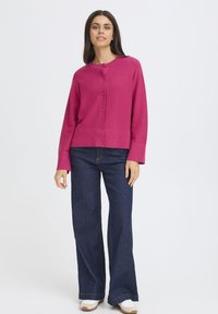 Cardigan tricotat fucsia cu gât rotund, închidere cu butoane în față și mâneci lungi, asortat cu blugi largi de culoare albastru închis și adidași albi.