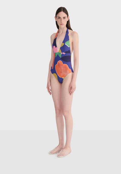 Femme debout portant un maillot de bain une pièce à encolure en V profonde avec des motifs floraux abstraits colorés et des chaussures beige tissées sans lacets.