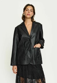 Zwarte leren blazer met een gesneden kraag, twee frontzakken en knoopsluiting. Soepele textuur en losse pasvorm. Ggedragen over een doorschijnende kanten rok.