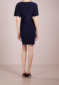 Kvinna som står vänd bortåt och bär en marinstocksblå knälång klänning med vita sidopaneler och svarta högklackade skor.