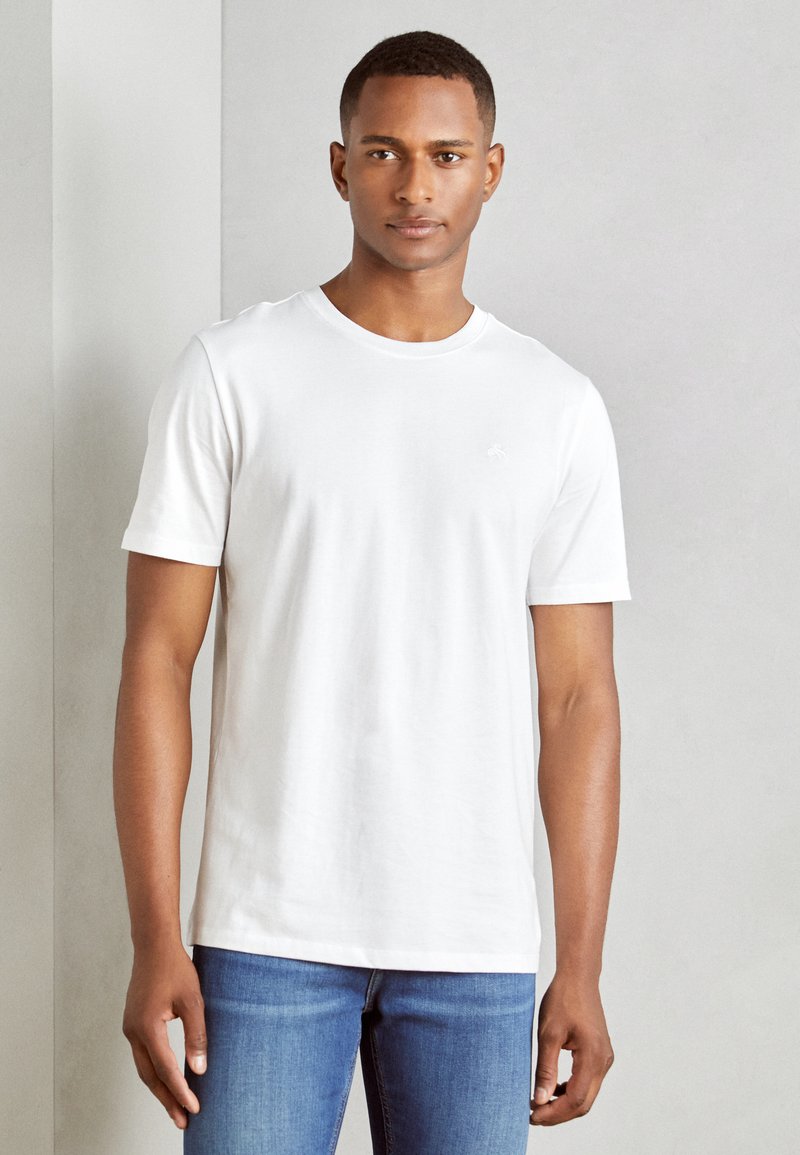 White cotton t-shirt, crew neckline, short sleeves, relaxed fit, plain design with a small logo on the chest, paired with blue jeans.
