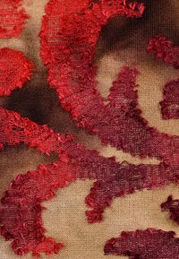 Textil mit komplexen fransigen Mustern in Rot und Burgunderrot auf einem hellbraunen Hintergrund, das ein strukturiertes Stoffdesign zeigt.