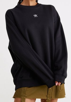 Person trägt einen übergroßen schwarzen Adidas-Sweatshirt mit Logo auf der Brust und einen braunen Rock unterhalb des Knies mit Riemendetails.