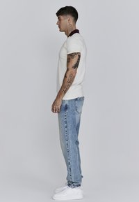 Witte polo met een kastanjebruine kraag, gecombineerd met lichtblauwe, versleten jeans. Model staat zijwaarts en draagt witte sneakers.