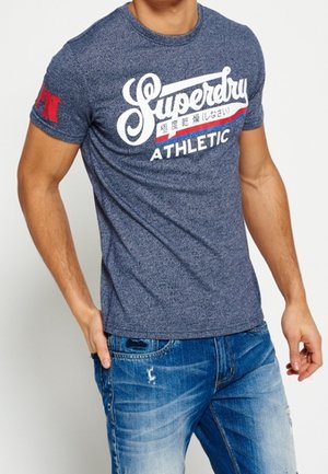 Homme portant un t-shirt bleu Superdry Athletic et un jean bleu délavé, une main dans la poche et l'autre détendue sur le côté.