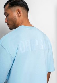 Lysblå oversized t-shirt lavet af blødt stof. Den har et tekstureret, tonalt "DROPSIZE" print på ryggen og en klassisk rund hals-design.