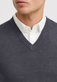 Kohlegrauer V-Ausschnitt-Pullover mit strukturiertem Strickdesign, kombiniert mit einem frisch weißen Hemd mit Kragen, das den Layering-Stil hervorhebt.