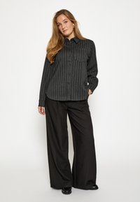 Camicia a righe nere e grigie con un'unica tasca sul petto, abbinata a pantaloni neri a gamba larga. Tessuto morbido, vestibilità su misura.