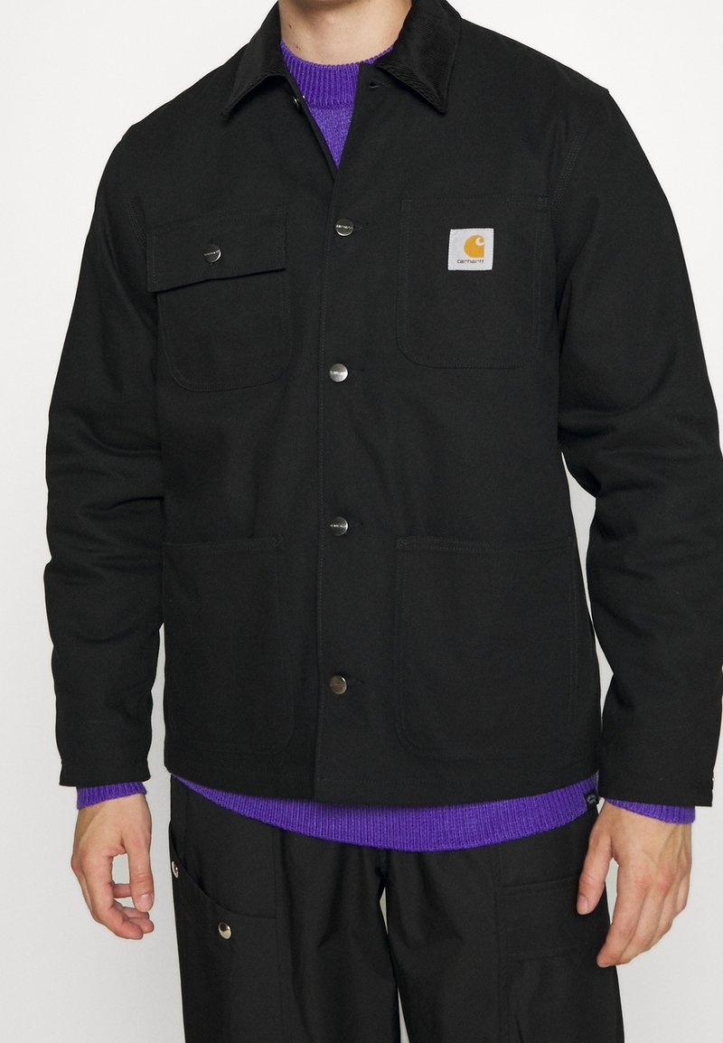 Giovane che indossa una giacca Carhartt nera con bottoni a pressione, tasche a toppa, sopra un maglione a coste viola e pantaloni neri abbinati.