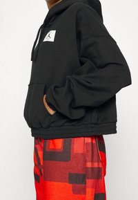 Felpa corta nera con tasca frontale, cappuccio regolabile con cordino e patch con logo. Abbinata a pantaloni rossi con motivo testurizzato.