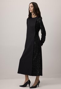 Largo vestido negro con un corpiño ajustado y mangas adornadas con lentejuelas. Presenta una silueta recta y lazos laterales. Tacones altos negros brillantes.