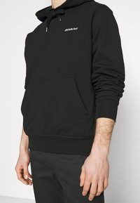 Osoba v černé mikině Dickies s klokankou a černých kalhotách, stojící proti jednoduchému světlému pozadí.