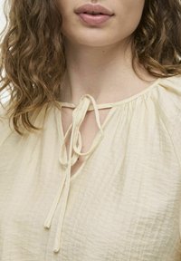 Blusa beige claro con un tejido texturizado. Presenta un cuello con un detalle de lazo en el escote y sutil fruncido en los hombros.