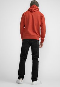 Felpa rossa con cappuccio, abbinata a jeans neri. L'outfit presenta un design semplice e una texture liscia, completato da sneakers bianche.