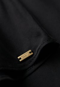 Czarny materiał o gładkiej strukturze. Zawiera małą złotą metalową metkę z wytłoczonym napisem "SUPER DRY", przymocowaną dwoma czarnymi nitkami.