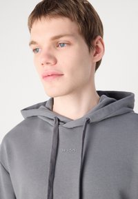 Grijze hoodie met trekkoord, voorzien van een subtiele logo-embossing op de borst, gemaakt van zacht materiaal met een gladde textuur.