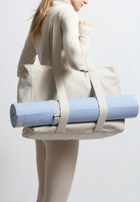 Plátěná taška v světle béžové barvě, s pevnými uchy, která drží sbalenou modrou jógovou podložku. Taška má texturovaný povrch a cylindrický tvar.