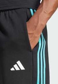 Pantalones deportivos negros con tres rayas verticales de color azul claro, un logo blanco de Adidas y un bolsillo, fabricados con un tejido suave y transpirable.