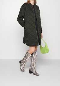 Pikowana zielona kurtka bomberka, czarna mini sukienka, wzorzyste kozaki do kolan oraz mała, usztywniana zielona torebka.