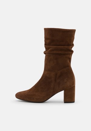 Brązowe zamszowe botki za kostkę o luźnym fasonie, spiczastym nosku i grubym obcasie. Widać gładką teksturę i proste detale przeszyć.
