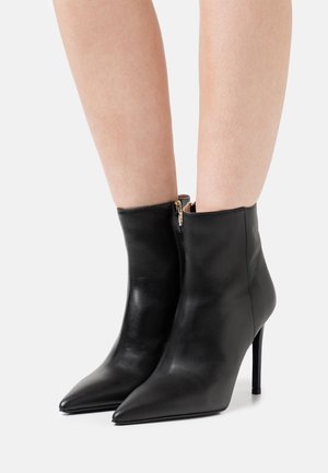 Botines de cuero negro hasta el tobillo con punta afilada, forma estrecha y cremallera lateral. Presentan un tacón alto delgado y una textura suave.