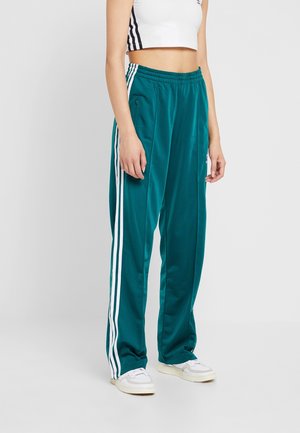 Kobieta ubrana w turkusowe spodnie dresowe z białymi paskami i białe trampki, w połączeniu z białym skróconym topem na ramiączkach z czarnymi paskami.