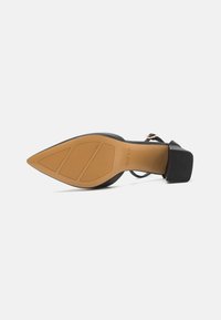 Black heeled sandal with a pointed toe, textured upper, and a rubber sole featuring a patterned grip. Adjustable ankle strap included.