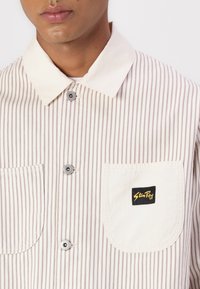 Striped button-up shirt with beige and light brown stripes, pointed collar, metal buttons, and a chest pocket displaying a black and yellow logo.