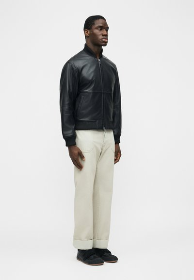 Άνδρας μοντέλο όρθιος, φοράει μαύρο δερμάτινο μπουφάν, μπεζ παντελόνι με φαρδιά μπατζάκια και ρεβέρ, και μαύρα παπούτσια, σε λευκό απλό φόντο.